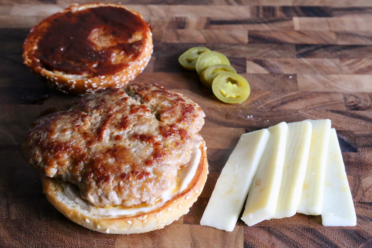 Italian Sausage Burger Assembly: toasted sesame buns, pork patty, garlic aioli, ketchup, mustard, pickled peppers and mozzarella.